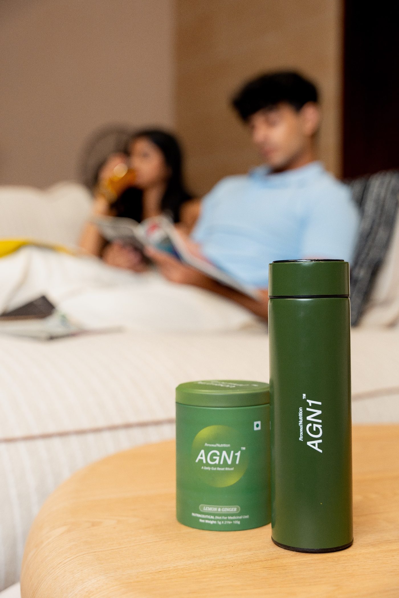 AGN1 supplement tub and green flask on a wooden table, with two people reading on a bed behind