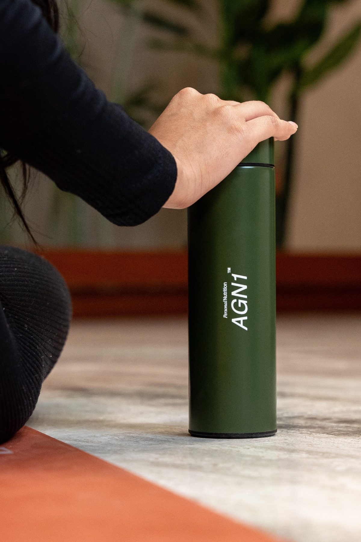Hand resting on a green AGN1 flask beside a person sitting on a rug