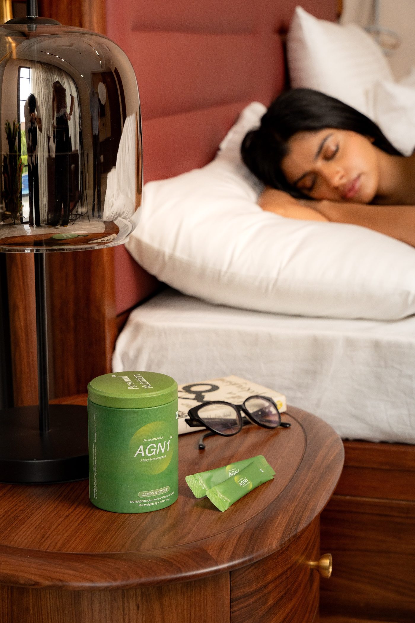 AGN1 tub and sachets on a wooden bedside table, with a woman asleep in the background