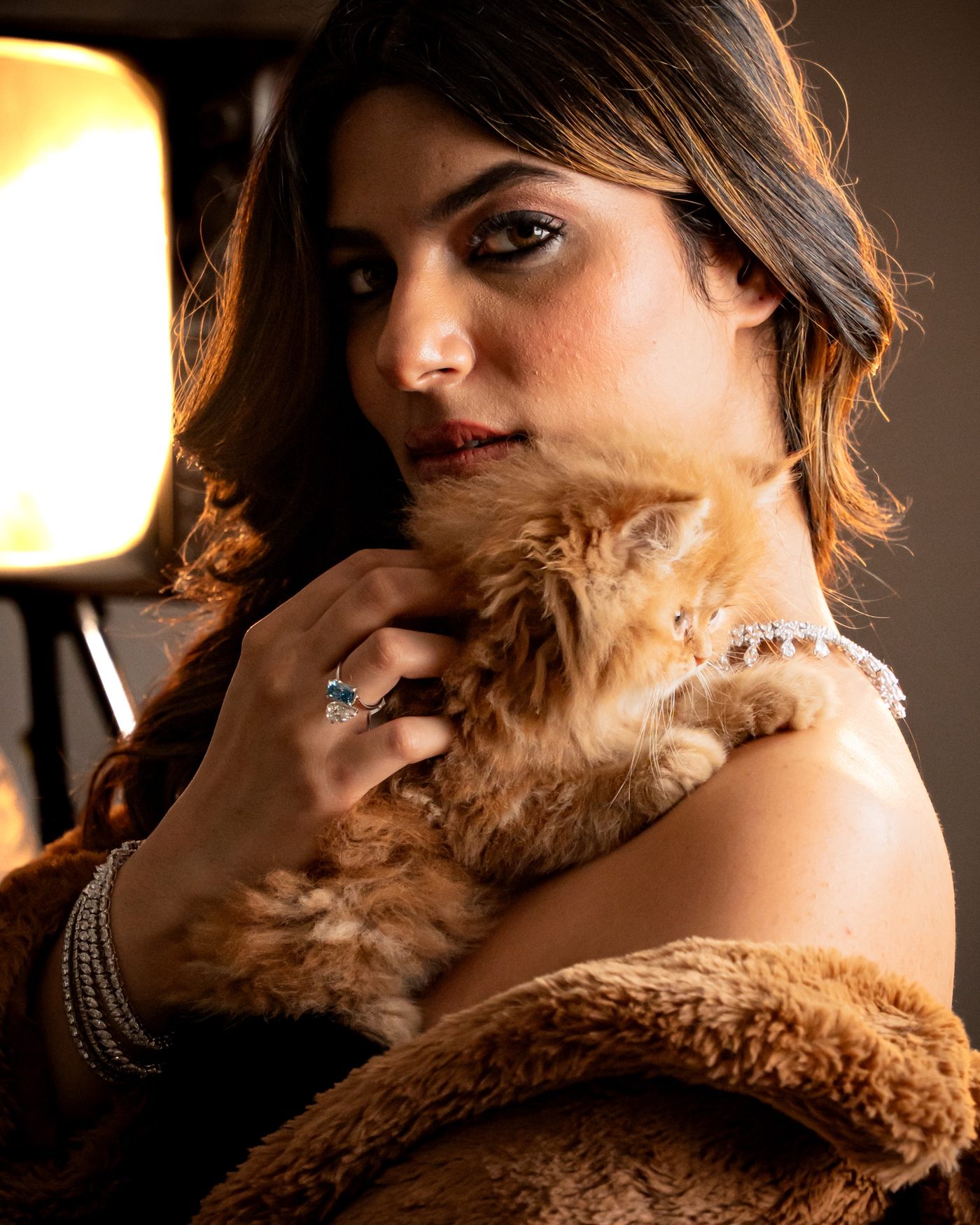Woman in a brown fur coat holding an orange kitten, wearing rings, bracelets and a necklace