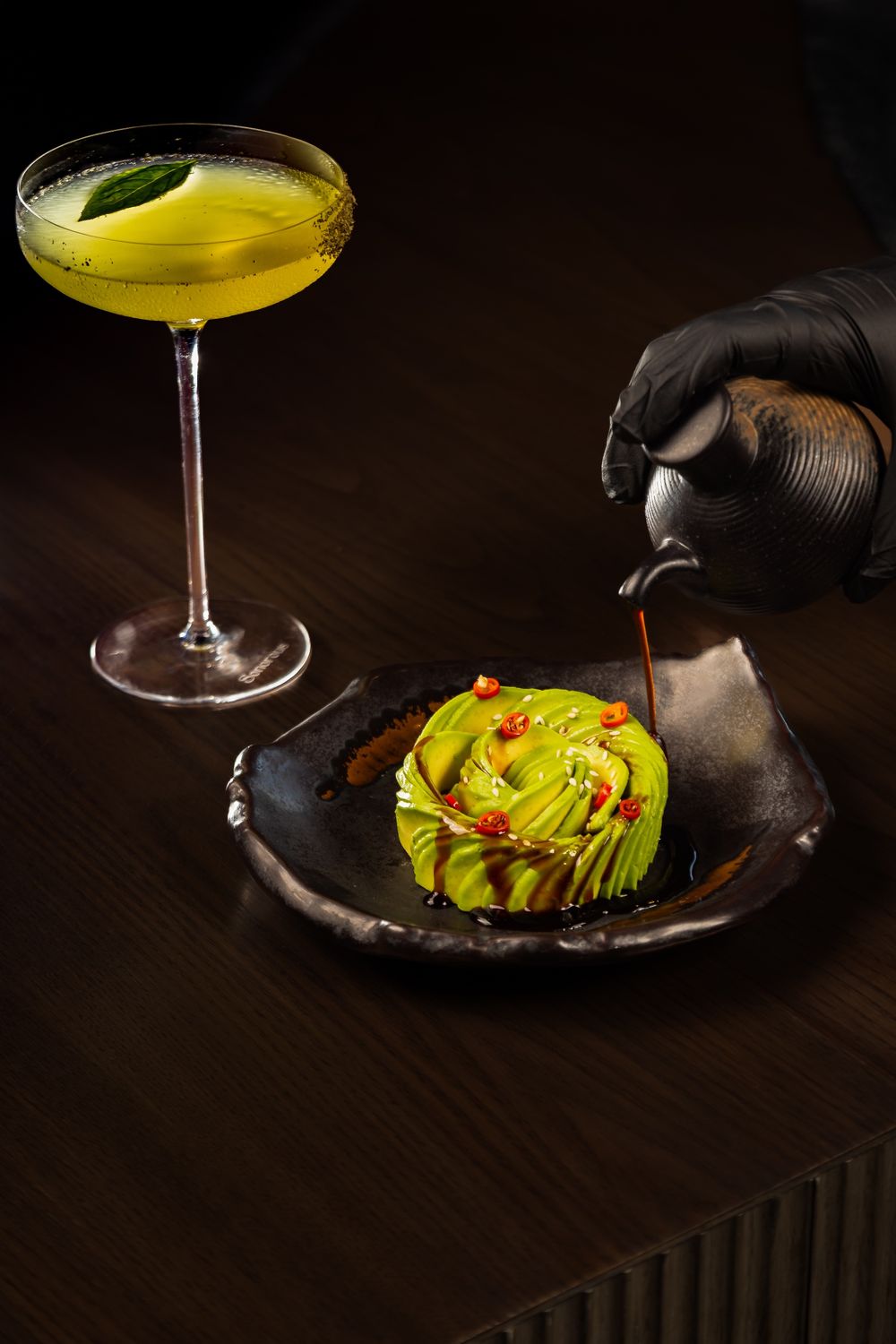 Green cocktail beside a sculpted avocado rose on a black plate as sauce is poured