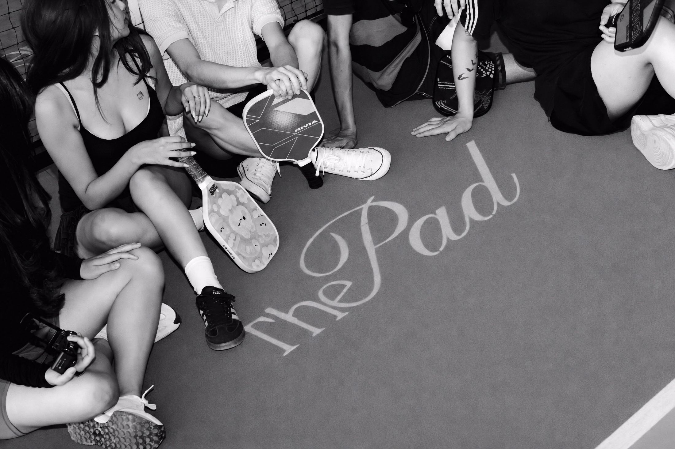 Players seated courtside around The Pad lettering on the court floor, in black and white